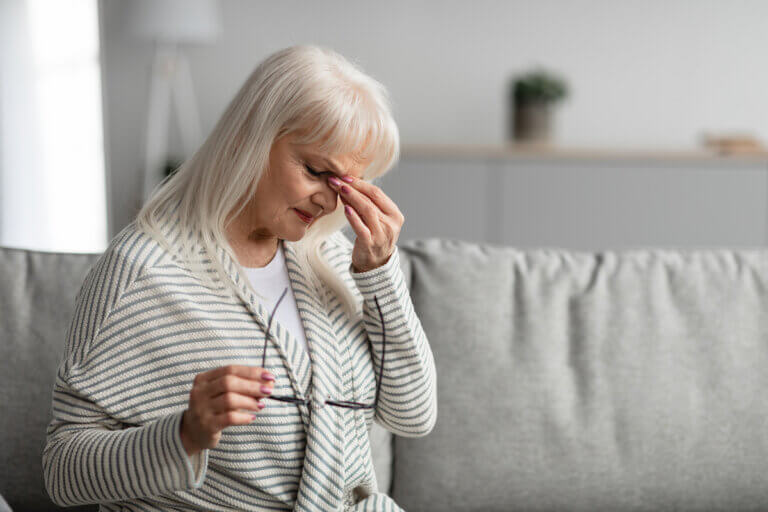 Portrait of tired mature woman massaging her nosebridge, rubbing dry irritated eyes sitting on the couch at home, free copy space. Female exhausted from looking at screen, holding eyeglasses